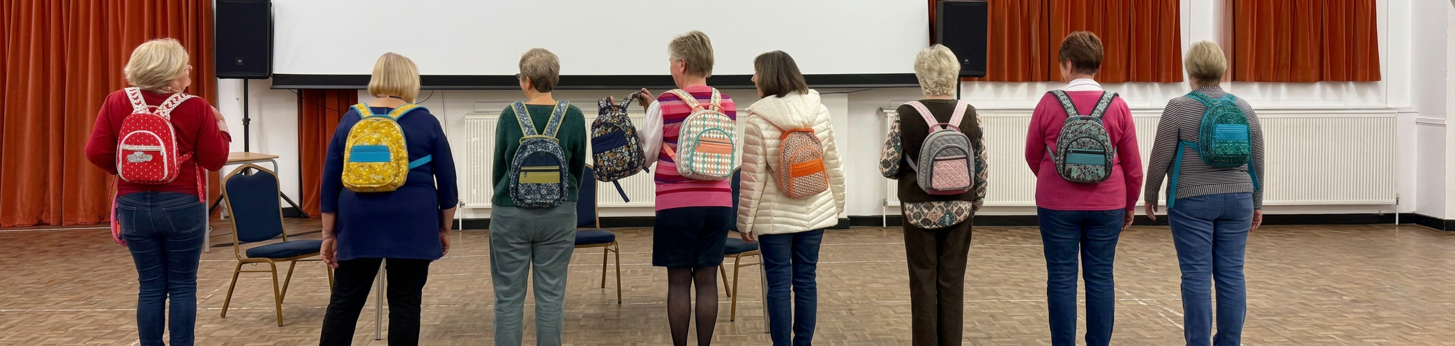 A group of eight women stands with their backs to the camera, each wearing colorful backpacks. They are positioned in a line in front of a blank screen in a spacious room with orange curtains.