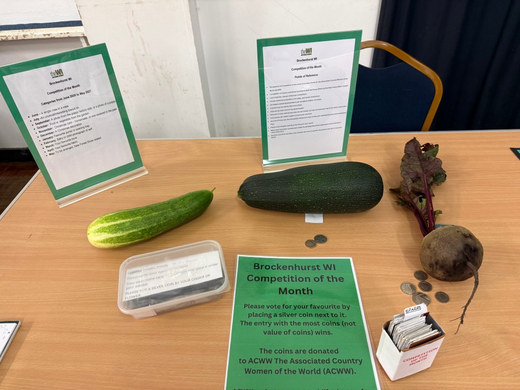 Display of vegetables for the Brockenhurst WI Competition of the Month, featuring a cucumber, a zucchini, a beetroot, and a voting box with coins on a table.