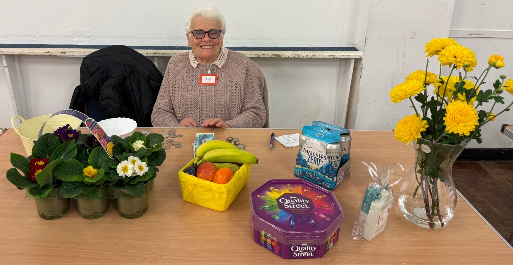 An elderly woman with glasses smiles behind a table filled with potted flowers, fruit, a box of Quality Street chocolates, and other items.