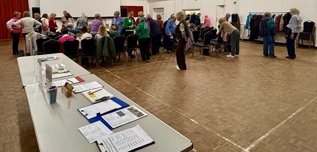 A group of people, mostly older adults, mingling and standing around in a community hall. Some are seated at tables with papers and supplies, while others are engaged in conversation. The room is spacious with chairs arranged in rows and clothes hanging on a rack in the background.